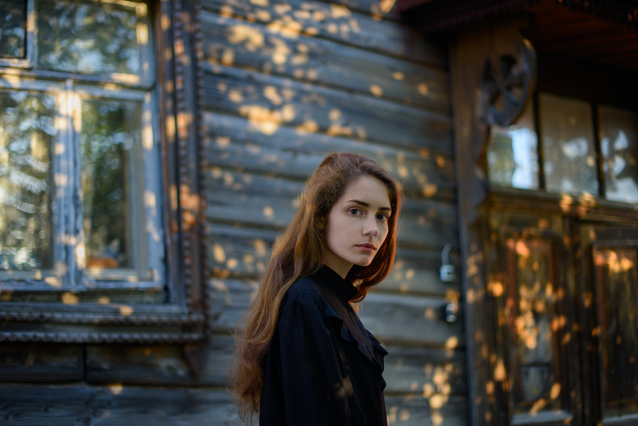 Young woman with long hair in front of a wooden house in dappled evening light