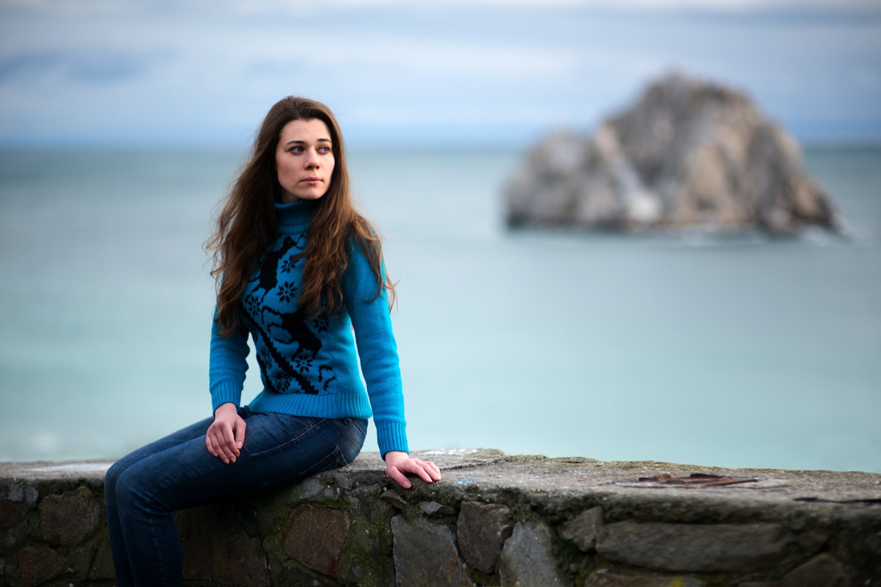 Woman in a blue sweater sitting on a stone wall by the sea, framed wider