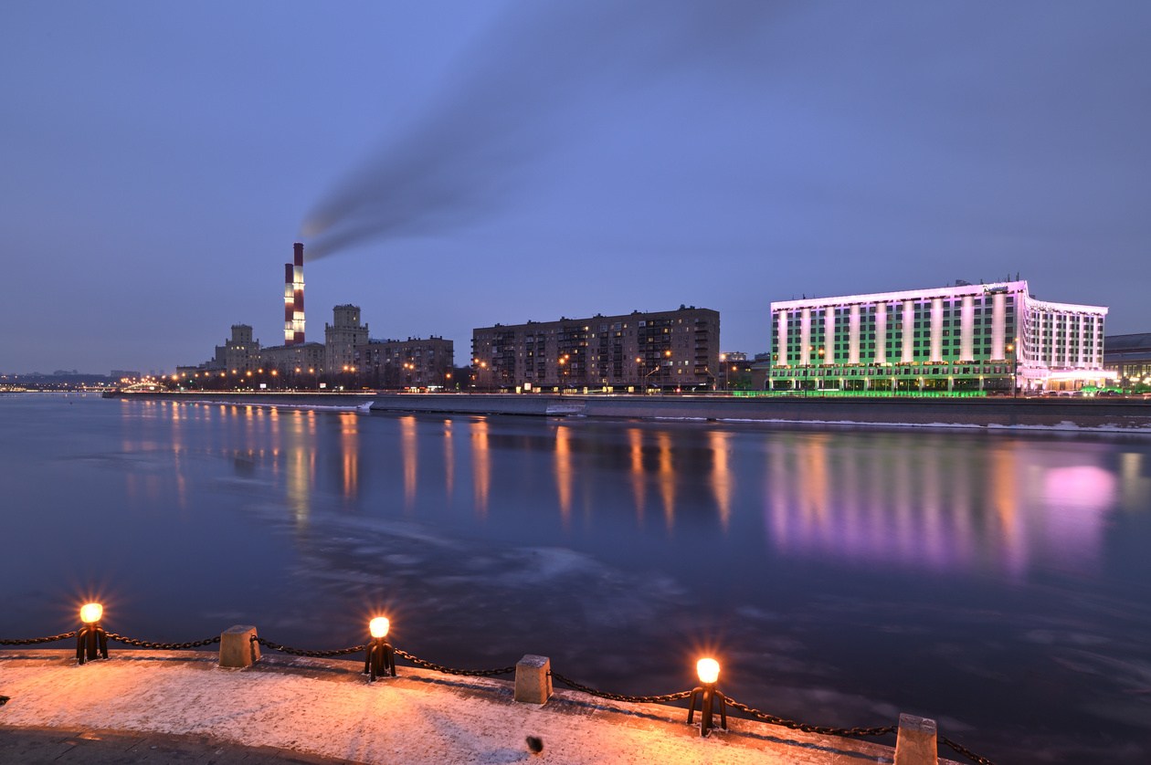 River embankment at blue hour with a power-station chimney and lit buildings reflected in the water