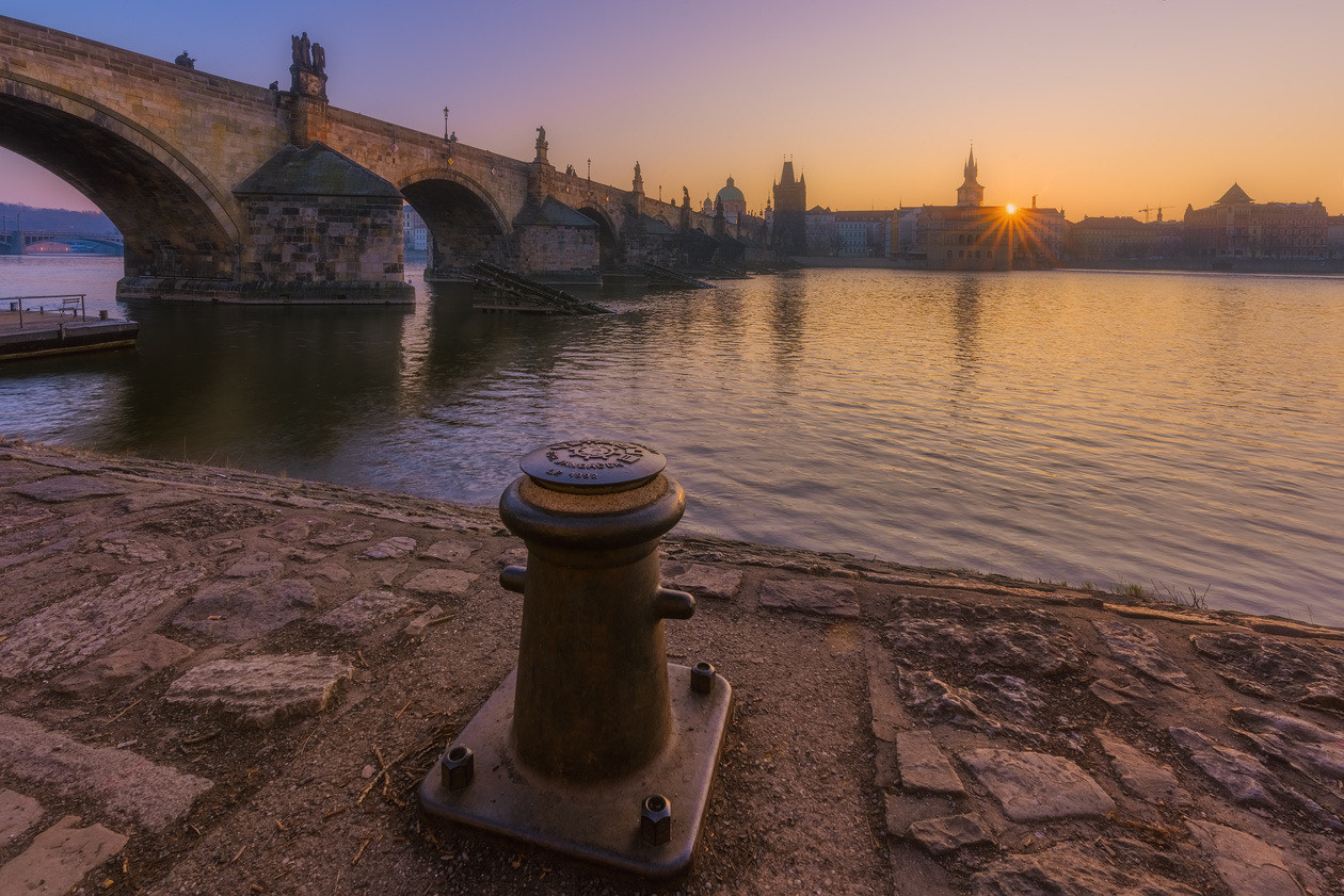 Riverbank at sunset with a stone bridge and a mooring bollard in the foreground