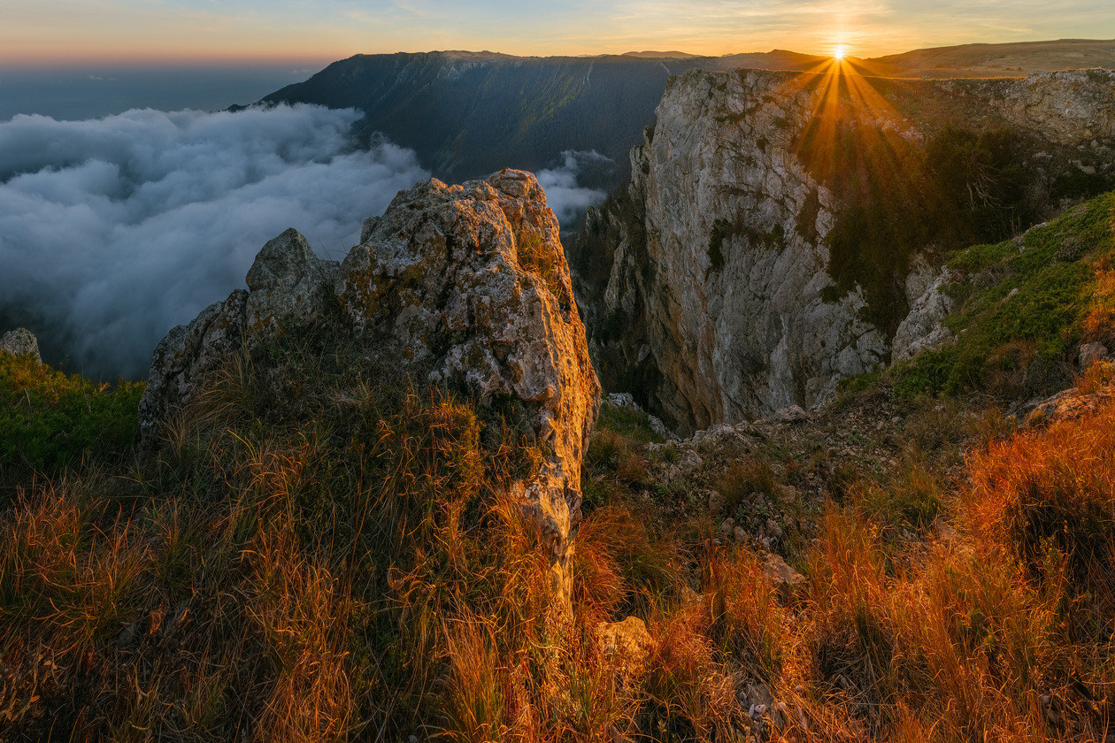 Rocky cliffs above a sea of clouds with the sun on the horizon