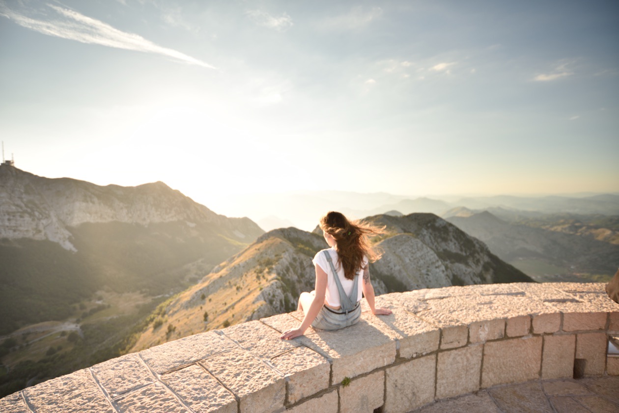 Young woman sitting on a stone parapet facing mountains under a bright hazy sky