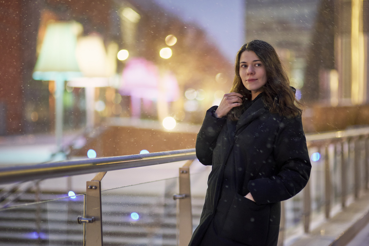 Young woman in a dark coat on a snowy evening embankment with soft city lights behind her
