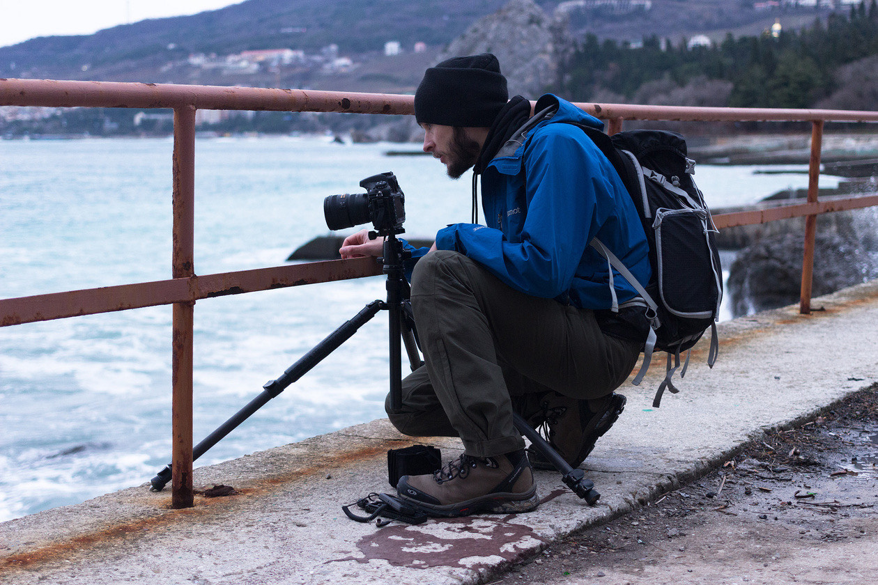 Photographer crouching behind a camera on a tripod at a seaside railing