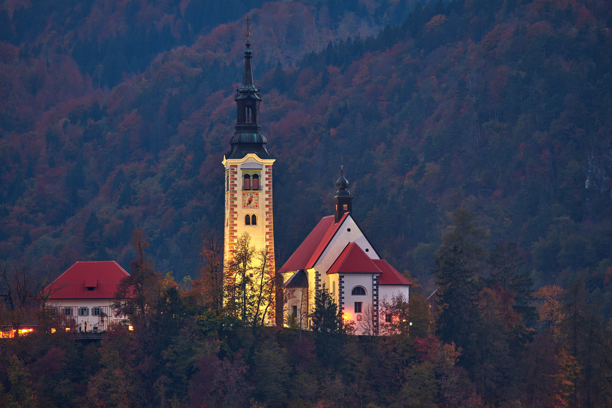 Lit church on a wooded hill at dusk with autumn trees around it