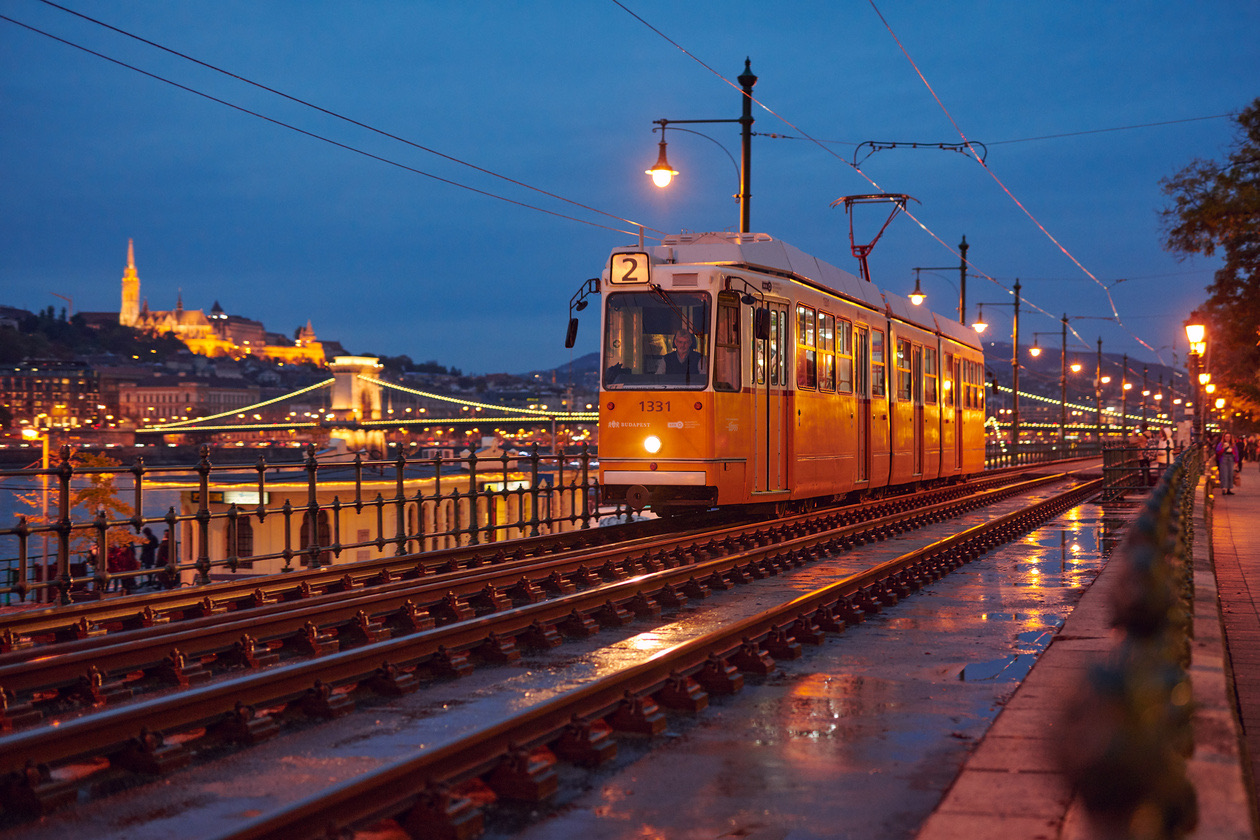 Yellow tram on rain-wet tracks along an embankment at dusk with a castle behind