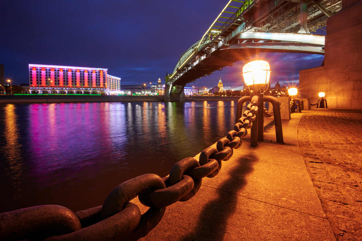 Riverside promenade at night with a heavy chain, a glowing lamp and a lit bridge