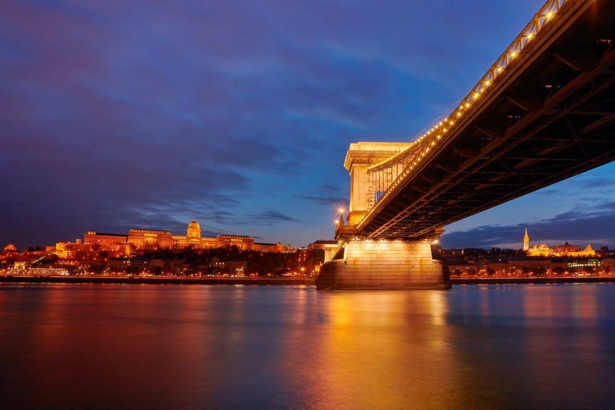 Illuminated chain bridge over a river at dusk with a deep blue sky