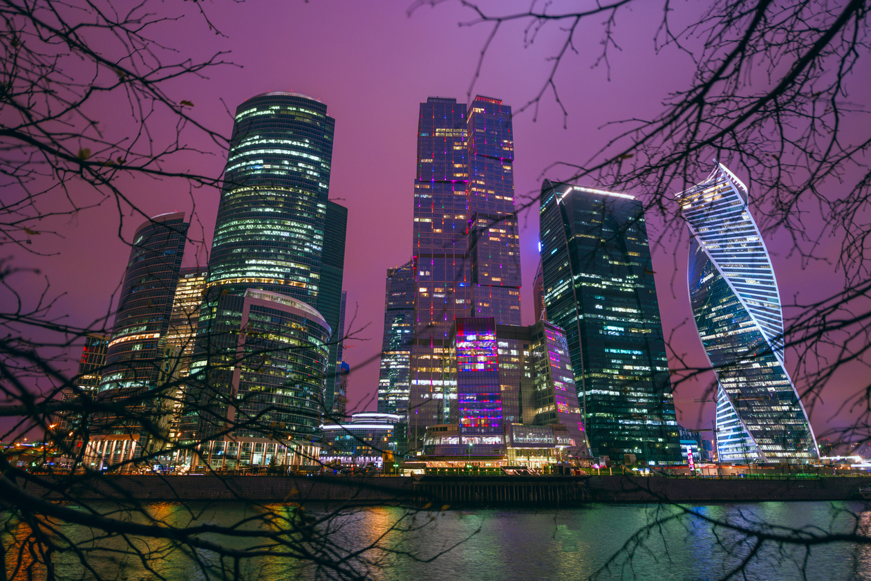 Glass skyscrapers lit up at night across a river behind bare branches