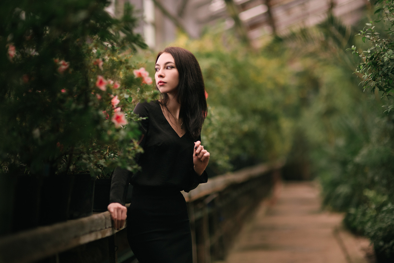Woman in a black dress in a greenhouse corridor surrounded by plants and pink flowers
