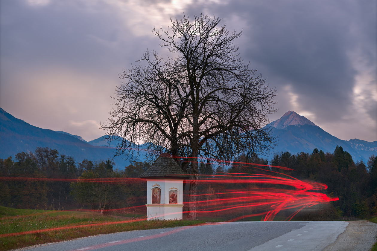 Red light trails from car headlights passing a small roadside chapel and a bare tree at dusk